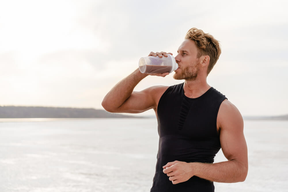 Muž v černém tílku pije proteinový nápoj u pláže